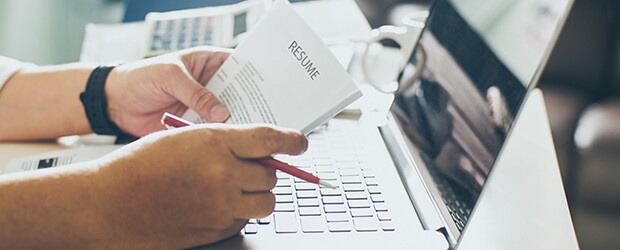 Hands holding a resume and a red pen while resting on a desk with a laptop