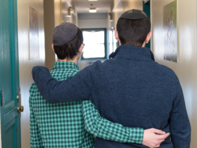 Two male Yeshiva University students walk down a hallway with their arms around their backs.