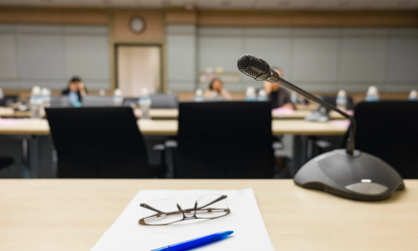 Glasses, pen and paper placed on a table in a lecture hall. There is a small mic to the right side of paper & pen.