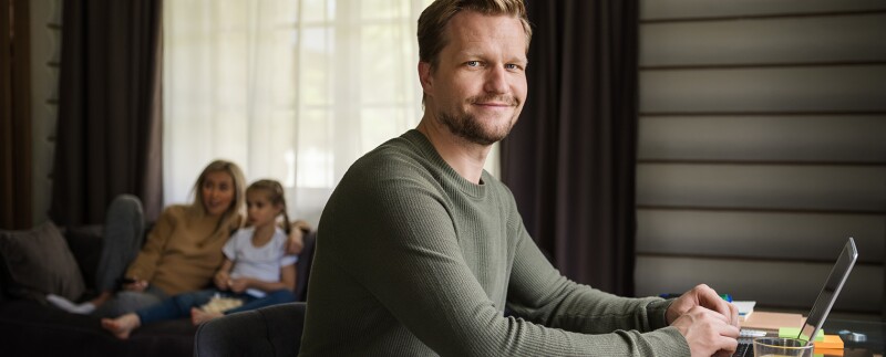 Smiling man working on laptop at a desk. His wife and daughter are watching tv on sofa in background