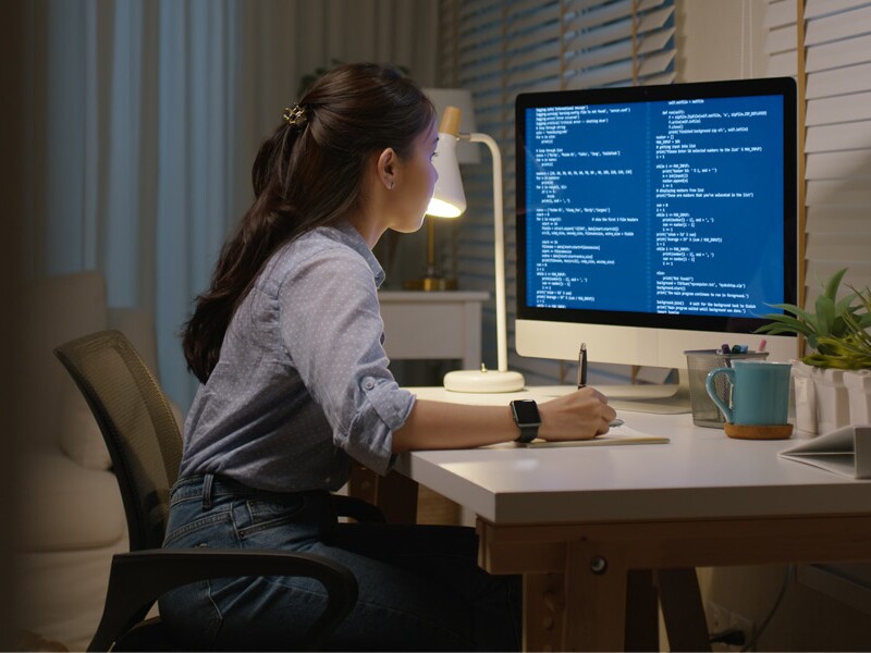 Young woman sits at a desk, studying a computer screen and taking notes.