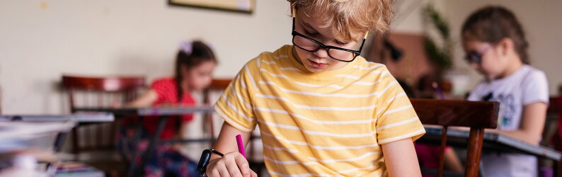 Blonde boy with glasses writing at a desk in class with classmates in the background