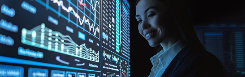 Young female data analyst reviews analytic metrics on touch screen wall monitor.