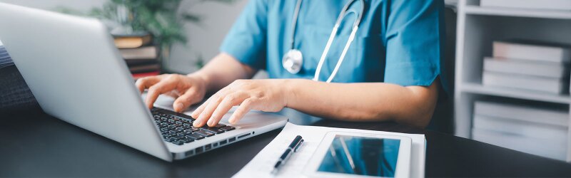 A nurse in scrubs typing on a laptop with a stethoscope around their neck, medical documents, and a tablet on the desk.