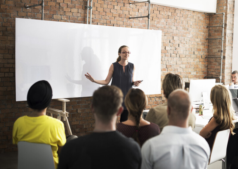 Woman standing in front of a white board leads a training session for a group of seated attendees