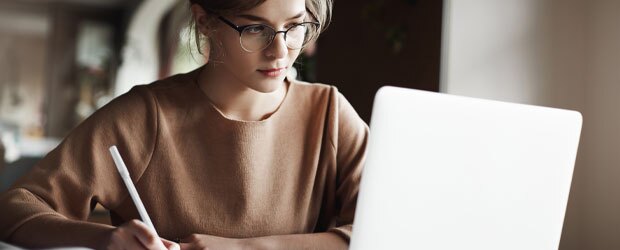Creative good-looking european female with fair hair is making notes while looking at laptop screen.