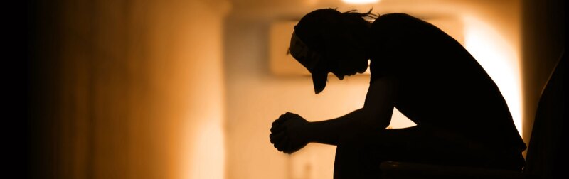 Silhouette of a young man sitting in a dark hallway with his arms crossed on his knees.