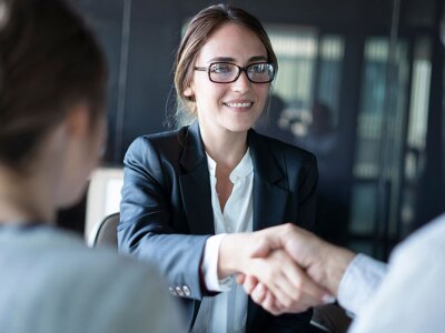 A woman in glasses and a suit smiles, shaking hands with someone across a table, potentially getting the job.