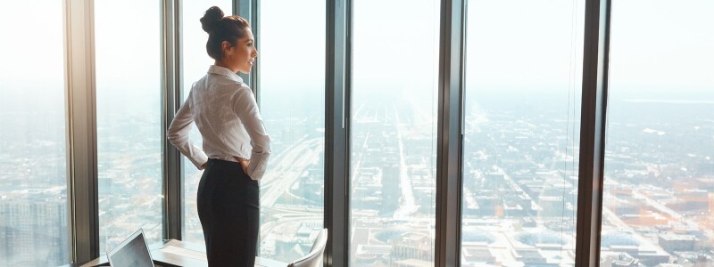 Woman standing in front of windows in a skyscraper with hands are on her hips admiring the view.
