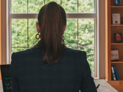 Woman sits at computer with her back to the camera. She's in front of a window in what seems to be a bookstore or a library.