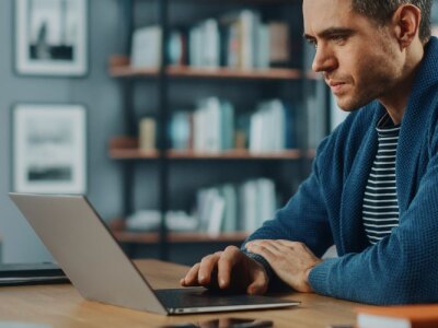 Male manager sits on a laptop at the kitchen table while communicating with team.