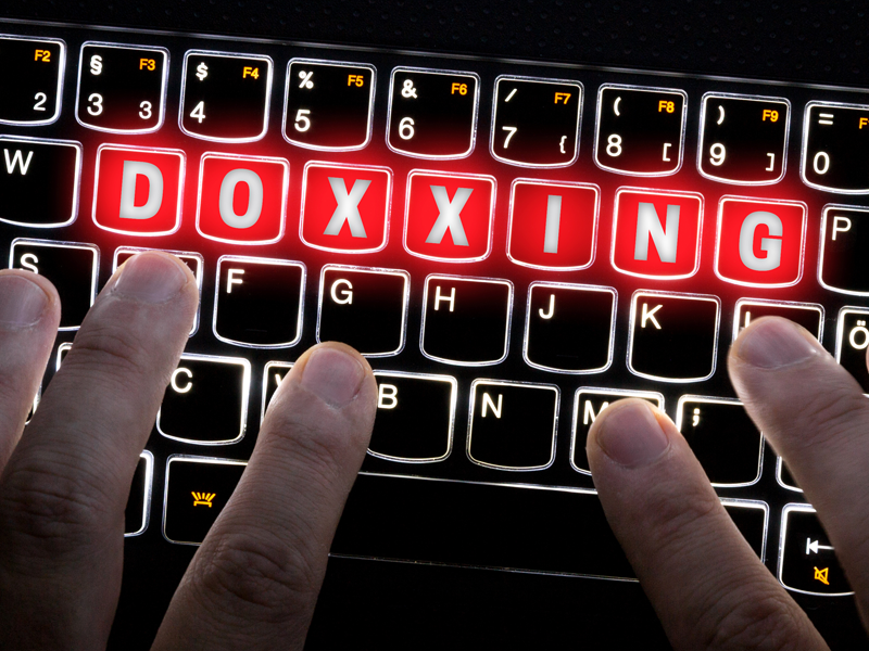 Hands are poised over the keys of a glowing keyboard. The word "doxxing" is highlighted on glowing red keys.
