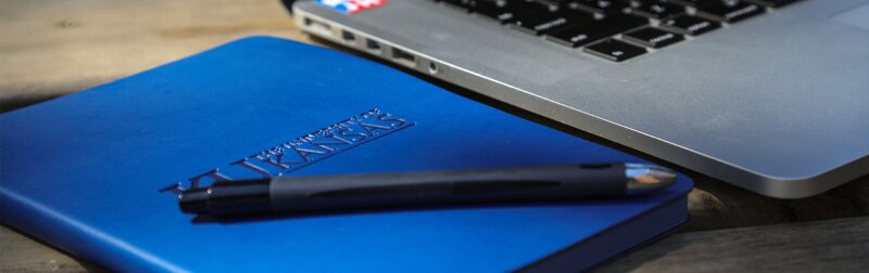 A close-up of a pen on a branded KU journal sitting next to a laptop.