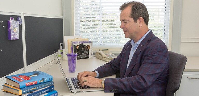 Man in blue blazer works on computer next to a pile of books