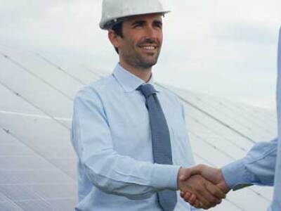Two businessmen, one in a hard-hat, shake hands in front of a bank of solar panels.