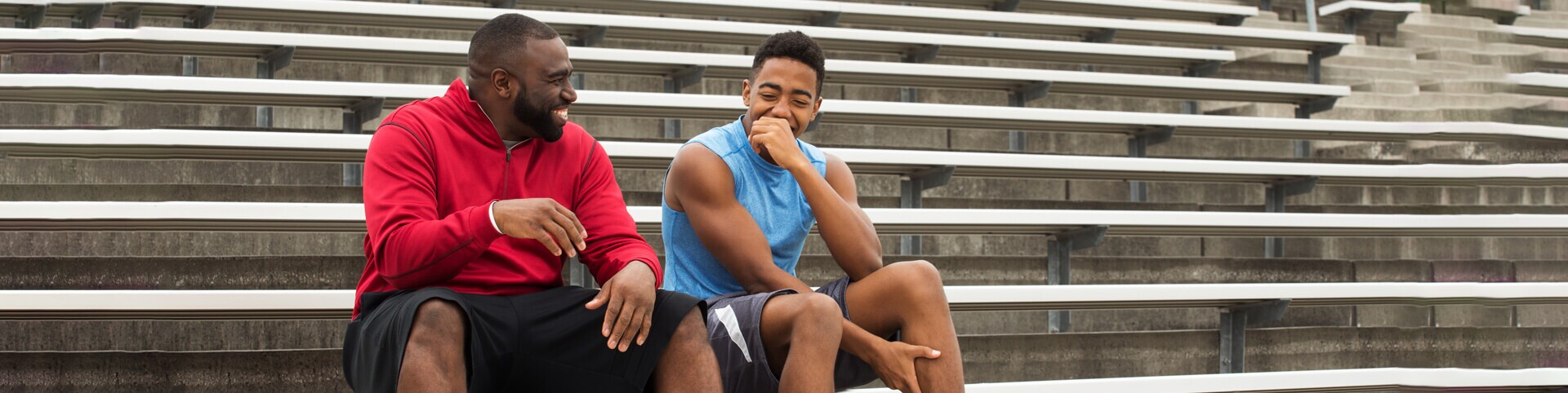 Two men in athletic clothing laughing on bleachers