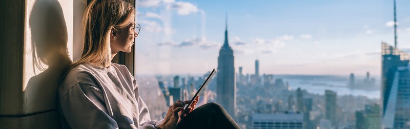 Woman with tablet looking out of a window at New York city