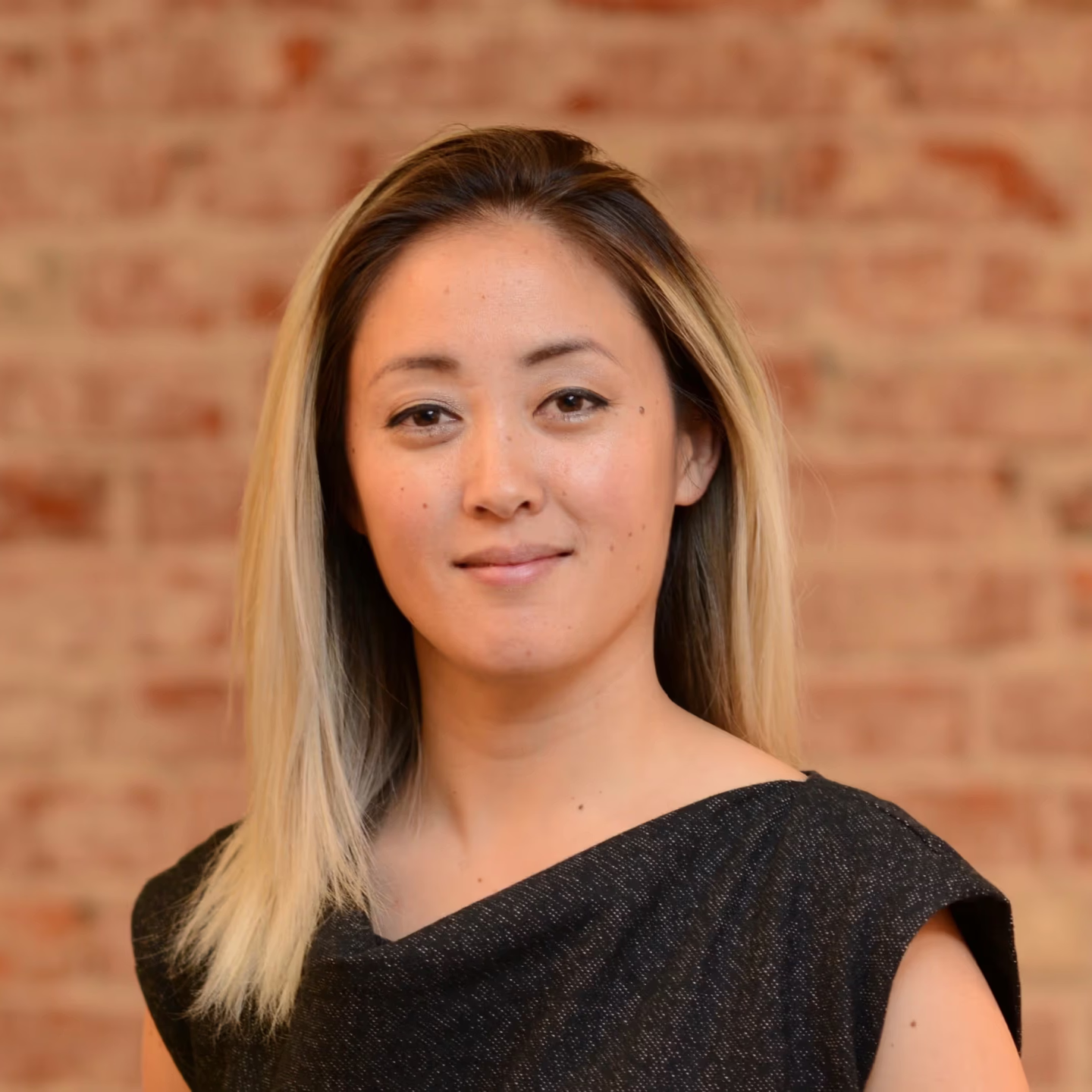 Professional headshot of a woman with shoulder-length dark-to-blonde hair, wearing a black top, standing against a warm brick background.