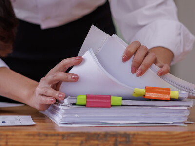 Close up shot of a woman's hands flipping through a thick stack of papers on a wooden desktop.