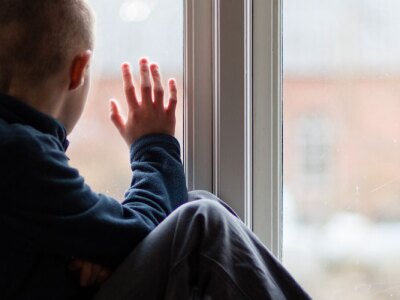 Young boy sitting on a windowsill looks out the window on a rainy day.