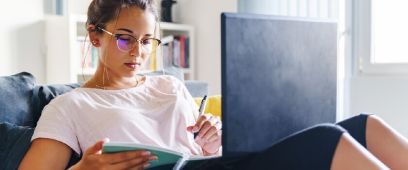 Young woman wearing glasses makes notes in notebook while working with laptop on her lap
