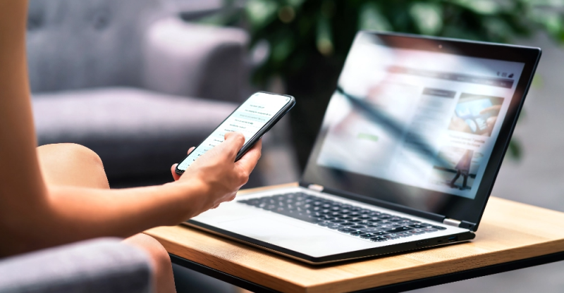 Female hands pictured holding a cell phone with a laptop in front of them.