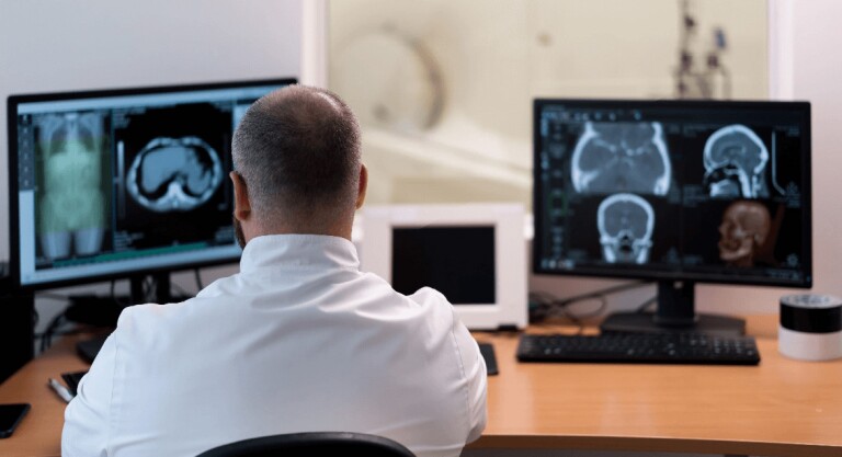 Over-the-shoulder view of a doctor seated at a desk examining brain and body scans