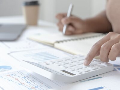 A person uses a calculator and writes in a notebook on a desk covered with financial charts, as well as a laptop. Going over accounting documents.