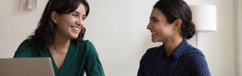 Two women having a conversation seated at desk in workplace.