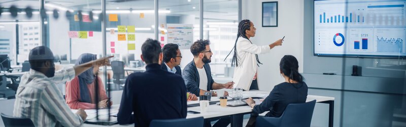 A diverse group of professionals in a marketing business meeting, a woman presents data on a large screen while colleagues observe and interact.
