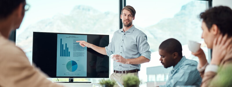 A man stands in a meeting room giving a presentation, pointing to a screen displaying charts and graphs, while several colleagues sit around a table listening and observing.