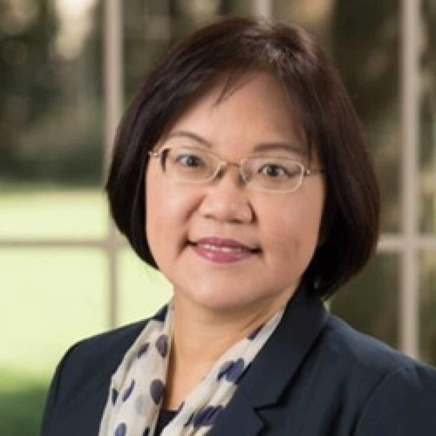 Professional headshot of a woman with short dark hair and glasses, wearing a blazer and patterned scarf, smiling in a softly blurred outdoor setting.