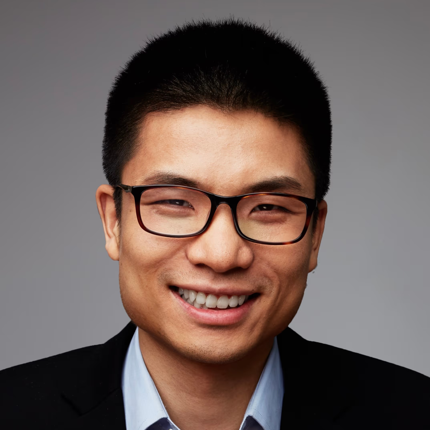 Headshot of a smiling man with short black hair and glasses, wearing a dark blazer and light shirt against a neutral background.