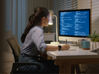 Young woman sits at a desk, studying a computer screen and taking notes.