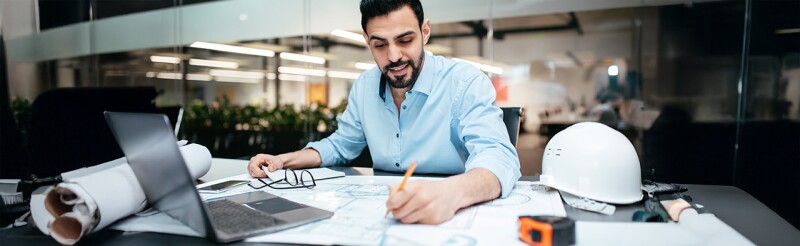 Engineer working on technical drawings with a laptop and safety helmet nearby