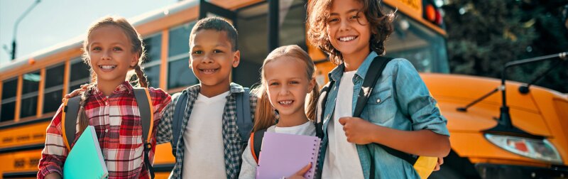 Group of school children stand together in front of a school bus.