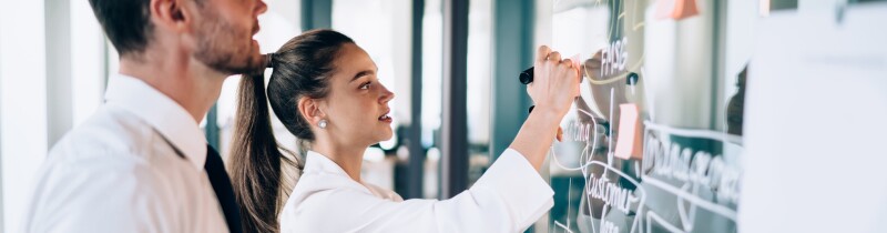 Woman writes with marker on glass board while man looks on.