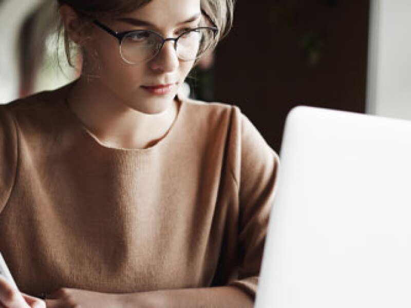 Creative good-looking european female with fair hair is making notes while looking at laptop screen.