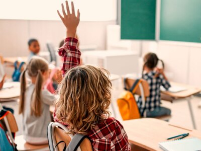 A classroom of students raise their hand to answer female teacher.