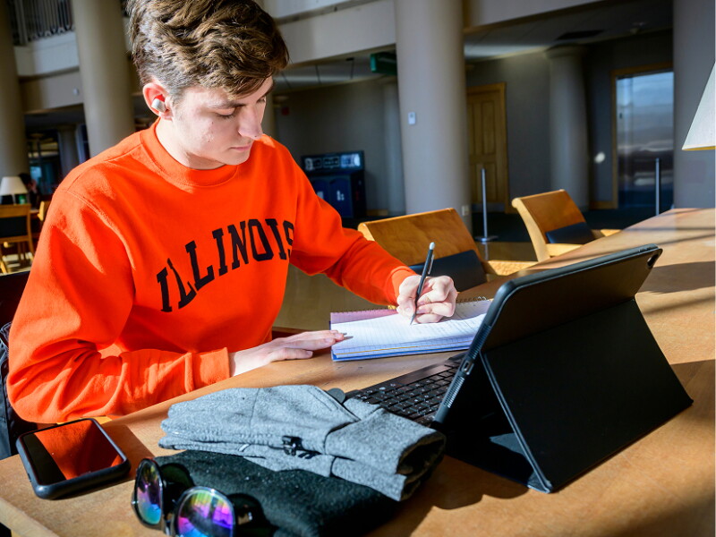 Male student wearing orange Illinois sweatshirt writing in notebook.