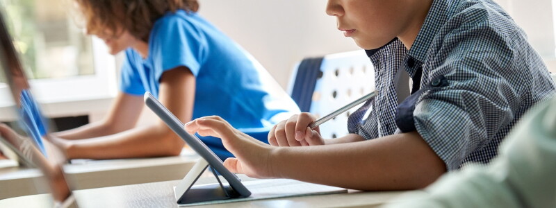Close up cut shot of Asian ethnicity elementary schoolboy using tablet device sitting in classroom with group of schoolchildren.