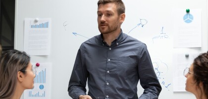Man leads a team meeting standing in front of a white boar
