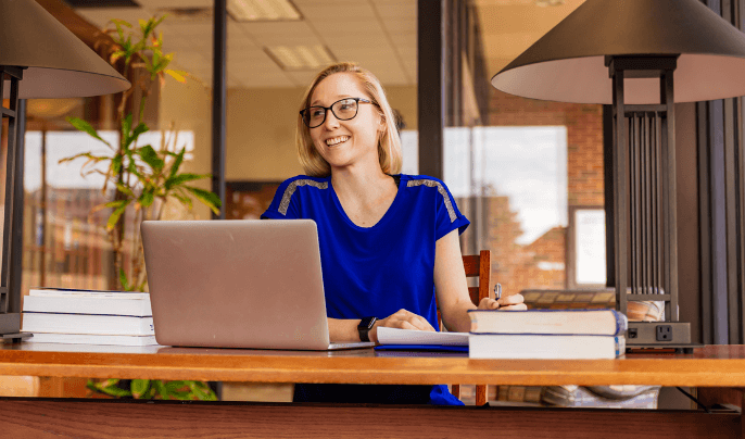 Woman in blue shirt seated at table writing notes and using laptop