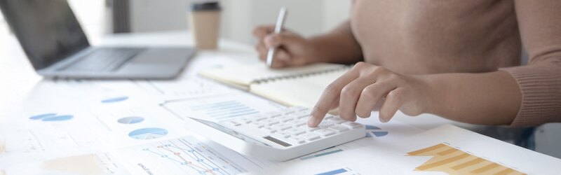 A person uses a calculator and writes in a notebook on a desk covered with financial charts, as well as a laptop. Going over accounting documents.