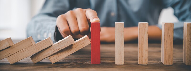 A man sitting at the table has his finger on top of the red block, stopping the remaining blocks from falling over.