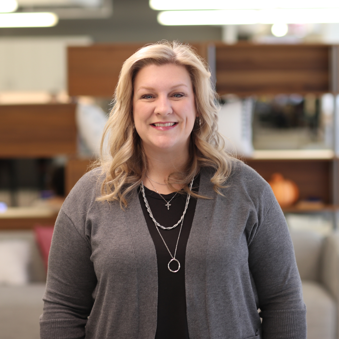 Professional headshot of Pam Stokes with long blonde hair, wearing a gray cardigan and layered necklaces in a modern office setting.
