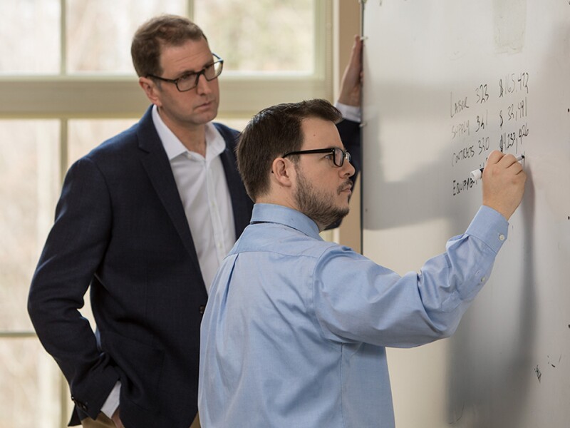 One man writing on a white board while another looks over