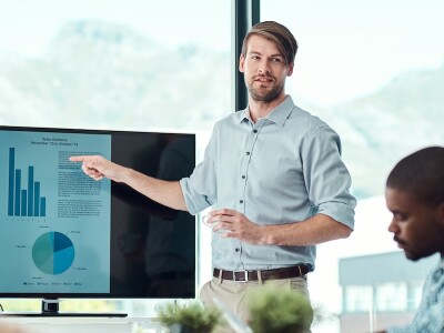 A man stands in a meeting room giving a presentation to a group, pointing to a screen displaying metrics.