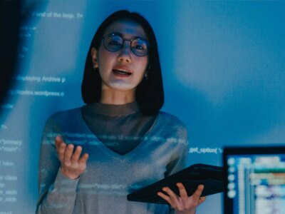 Woman speaking to others stands in front of a screen with code being projected onto it.