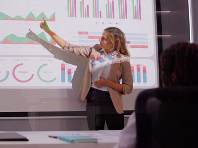 Business professional pointing to a screen displaying data charts and graphs in a meeting